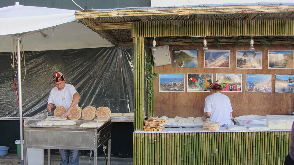 Baker in a traditional carapuça hat turning bolo do caco flatbreads on the griddle at a bamboo stall